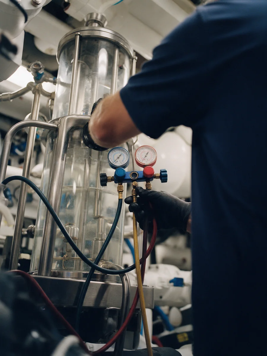 Gloved hands connecting a refrigeration gauge manifold to a chilled-water unit in a yacht machinery room